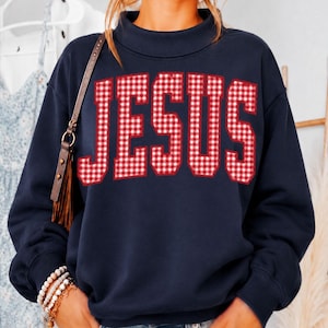 May include: A navy blue long-sleeve sweatshirt featuring a mock neck collar and the word JESUS printed across the chest in a red and white gingham check pattern. The garment has ribbed cuffs and a ribbed hem, providing a structured fit.