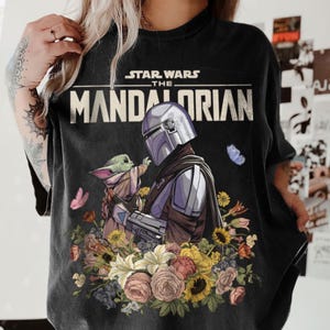 May include: Black t-shirt featuring a floral design with the Mandalorian and Grogu from Star Wars. The shirt has the text "STAR WARS THE MANDALORIAN" across the top. The design includes butterflies and flowers.