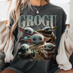 May include: Dark grey sweatshirt with the word "GROGU" in light blue above a graphic of five Grogu characters. The Grogu characters have light blue skin and large ears, in various poses.