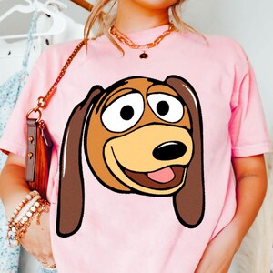 Pu&ograve; includere: T-shirt rosa chiaro con un'immagine a cartoni animati di Slinky Dog di Toy Story. La grafica mostra il volto di Slinky con orecchie marroni, un muso beige e un'espressione sorridente. La maglietta ha maniche corte e girocollo.