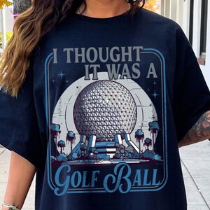 May include: Navy blue t-shirt with a graphic that reads "I thought it was a Golf Ball." The graphic features a geometric sphere design, with a blue and white color scheme. The shirt is a casual wear item.