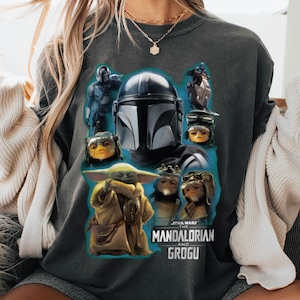 Puede incluir: Sudadera gris oscuro con un gr&aacute;fico de personajes de la serie The Mandalorian, incluyendo el Mandaloriano, Grogu y otras criaturas. El dise&ntilde;o est&aacute; sobre un fondo verde azulado, con las palabras "STAR WARS THE MANDALORIAN AND GROGU" impresas debajo.