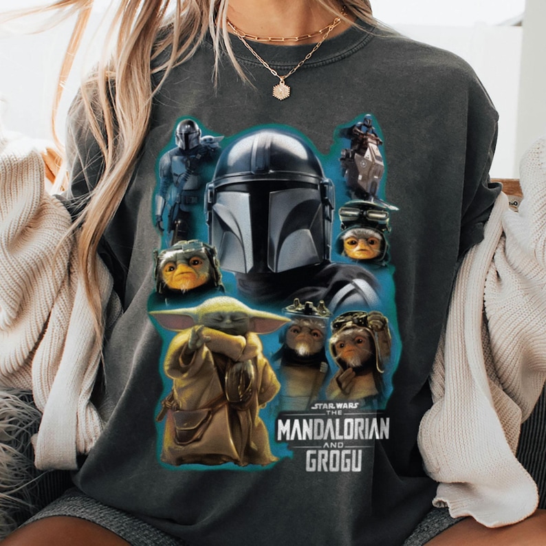 Puede incluir: Sudadera gris oscuro con un gr&aacute;fico de personajes de la serie The Mandalorian, incluyendo el Mandaloriano, Grogu y otras criaturas. El dise&ntilde;o est&aacute; sobre un fondo verde azulado, con las palabras "STAR WARS THE MANDALORIAN AND GROGU" impresas debajo.