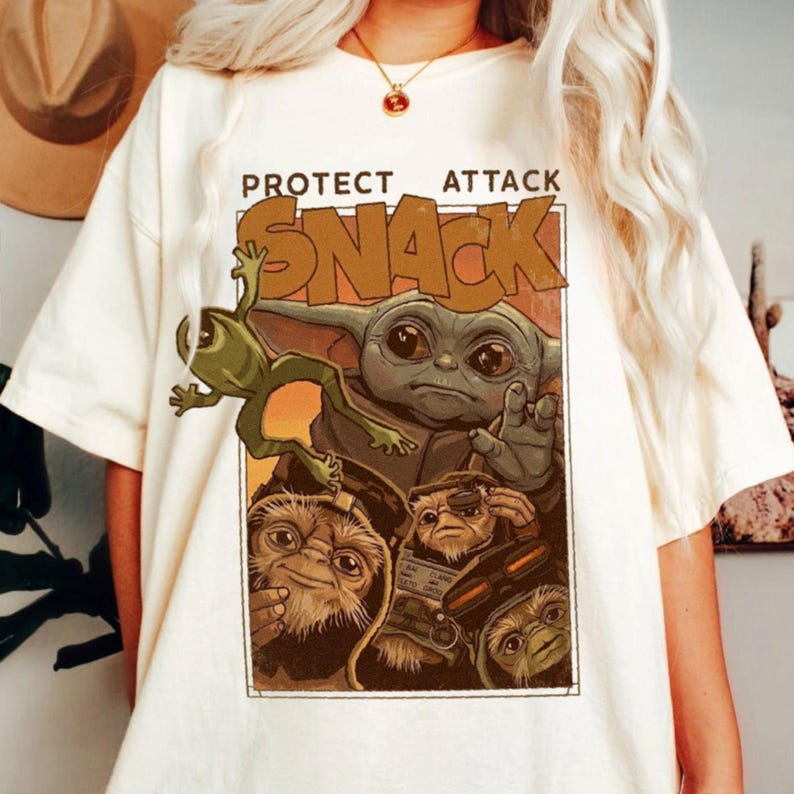May include: Cream-colored t-shirt featuring a graphic of a green frog-like creature, a gray alien-like character, and other characters. The text "PROTECT ATTACK SNACK" is printed above the characters in a brown font.
