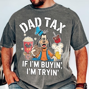 Op de afbeelding: Grijze t-shirt met de tekst "DAD TAX" en "IF I'M BUYIN', I'M TRYIN'". De t-shirt heeft een cartoon van Goofy met een pretzel, omringd door popcorn, een karamelappel, bier en een donut.