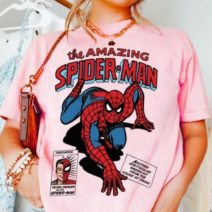 以下が含まれることがあります： スパイダーマンのコミックイラストが特徴のピンクのTシャツ。「the AMAZING SPIDER-MAN」の文字が赤と青でデザインされています。その他、コミック要素も含まれています。クラシックなスーパーヒーローをテーマにしたTシャツです。
