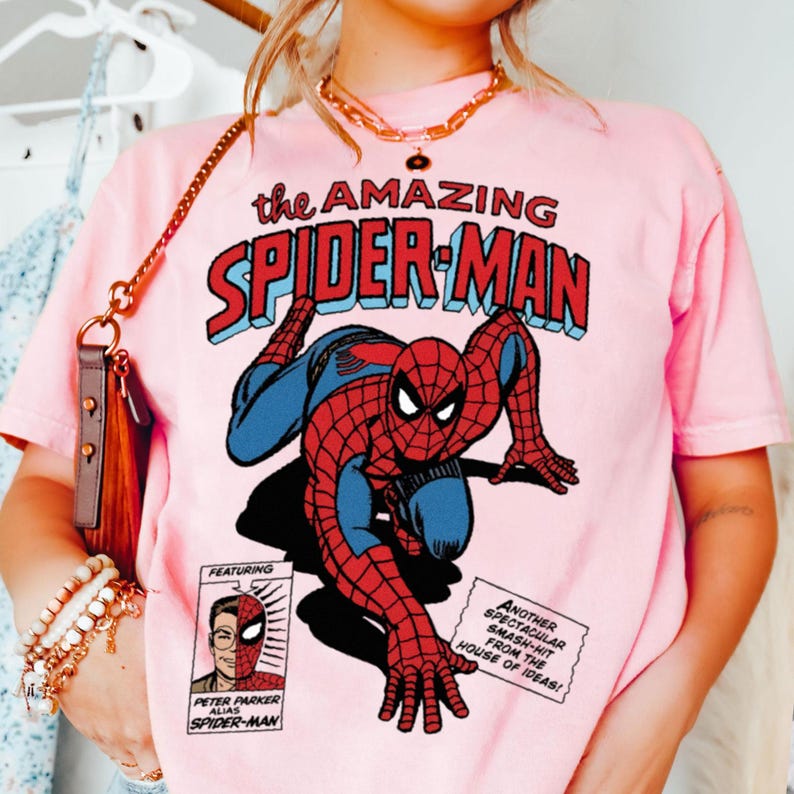 以下が含まれることがあります： スパイダーマンのコミックイラストが特徴のピンクのTシャツ。「the AMAZING SPIDER-MAN」の文字が赤と青でデザインされています。その他、コミック要素も含まれています。クラシックなスーパーヒーローをテーマにしたTシャツです。