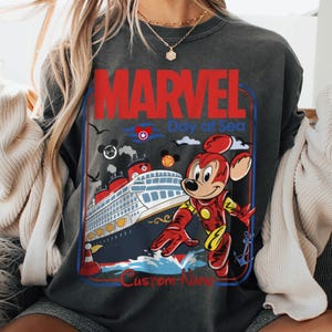 May include: Dark gray sweatshirt with a graphic featuring Mickey Mouse in an Iron Man suit, a cruise ship, and the words "MARVEL Day at Sea". The design includes red, blue, and yellow accents.