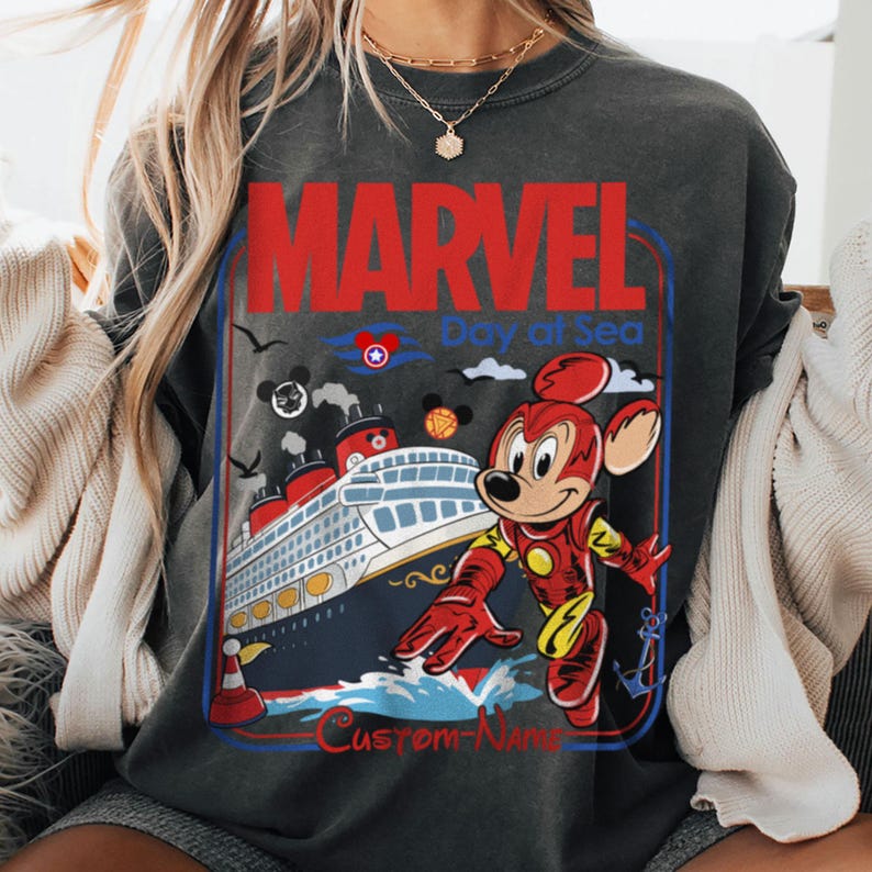 May include: Dark gray sweatshirt with a graphic featuring Mickey Mouse in an Iron Man suit, a cruise ship, and the words "MARVEL Day at Sea". The design includes red, blue, and yellow accents.