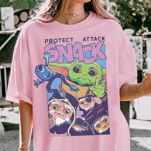 K&ouml;nnte beinhalten: Rosa kurz&auml;rmeliges T-Shirt mit einer Grafik eines gr&uuml;nen Aliens, eines blauen Aliens und anderer Charaktere mit dem Text "PROTECT ATTACK SNACK". Die Grafik hat eine rechteckige Form. Das Shirt ist aus weichem Material.
