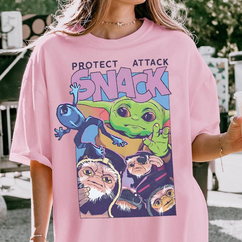 K&ouml;nnte beinhalten: Rosa kurz&auml;rmeliges T-Shirt mit einer Grafik eines gr&uuml;nen Aliens, eines blauen Aliens und anderer Charaktere mit dem Text "PROTECT ATTACK SNACK". Die Grafik hat eine rechteckige Form. Das Shirt ist aus weichem Material.