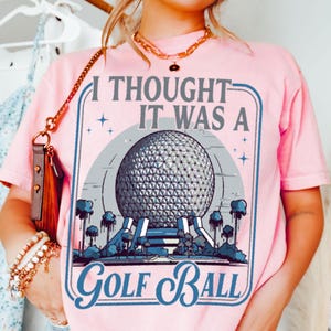 May include: Pink t-shirt with a graphic design featuring a large silver sphere, blue accents, and the text "I Thought It Was A Golf Ball." The design is framed in a blue border. The shirt has short sleeves.