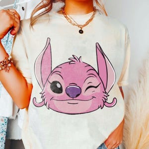 May include: Cream-colored t-shirt featuring a pink cartoon character with large ears and a winking expression. The character has a purple nose and a smiling mouth. The shirt is part of a casual wear collection.