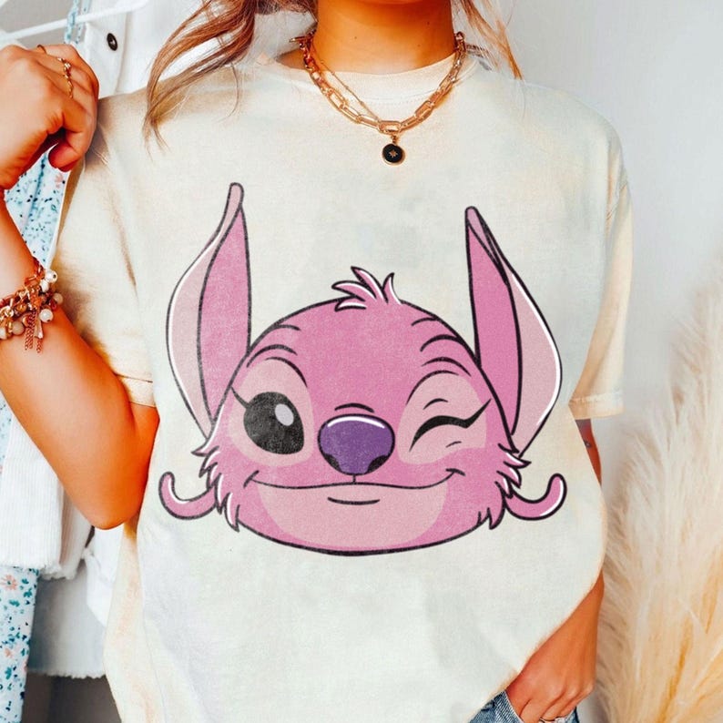May include: Cream-colored t-shirt featuring a pink cartoon character with large ears and a winking expression. The character has a purple nose and a smiling mouth. The shirt is part of a casual wear collection.