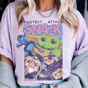 K&ouml;nnte beinhalten: Helllila T-Shirt mit einem Grafikdesign mit dem Text "PROTECT ATTACK SNACK". Die Grafik zeigt eine gr&uuml;ne, au&szlig;erirdisch aussehende Figur und andere Comicfiguren in einem farbenfrohen Design.