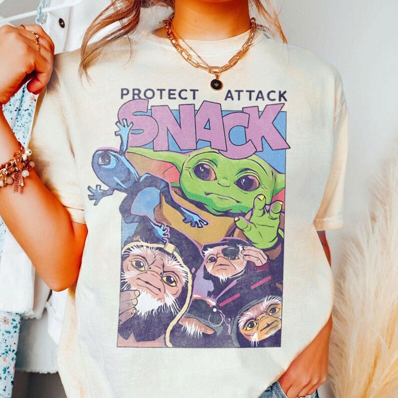 K&ouml;nnte beinhalten: Cremefarbenes T-Shirt mit einer farbenfrohen Grafik eines gr&uuml;nen Alien-Charakters, einer blauen Kreatur und anderen Charakteren. Der Text "PROTECT ATTACK SNACK" ist in Lila und Blau &uuml;ber den Charakteren aufgedruckt. Das Shirt hat kurze &Auml;rmel.