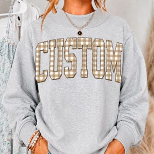 May include: A light gray crewneck sweatshirt featuring the word CUSTOM in large block letters across the chest. The letters have a tan and white gingham check pattern. A thin gold chain necklace with a small black circular pendant is worn.