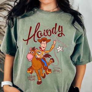 May include: Sage green t-shirt featuring a cartoon illustration of Woody from Toy Story riding Bullseye. The word "Howdy" is written in a burgundy script above the image. The shirt has a vintage, distressed look.