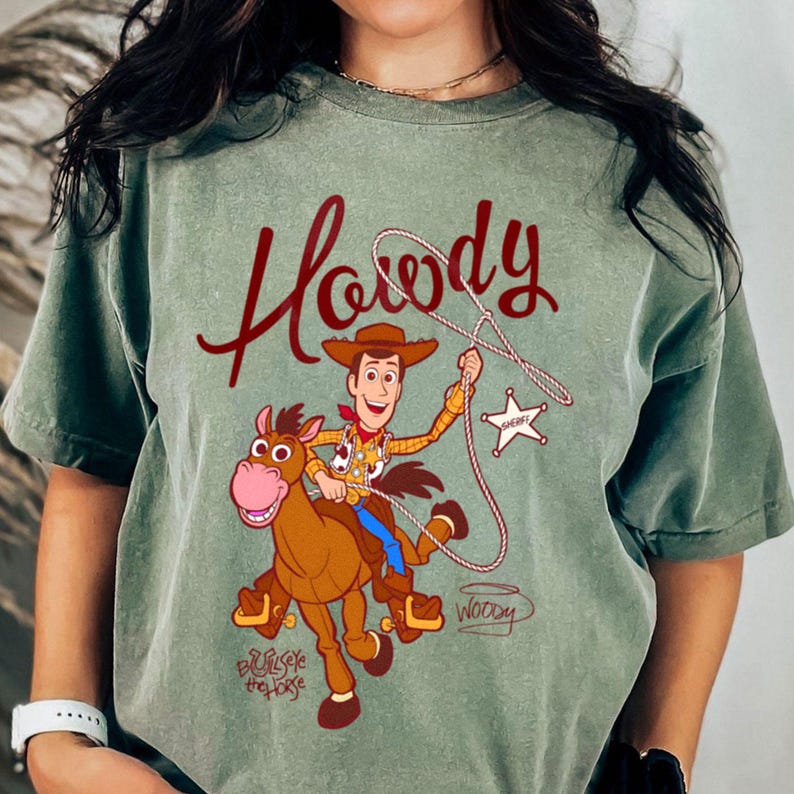 May include: Sage green t-shirt featuring a cartoon illustration of Woody from Toy Story riding Bullseye. The word "Howdy" is written in a burgundy script above the image. The shirt has a vintage, distressed look.