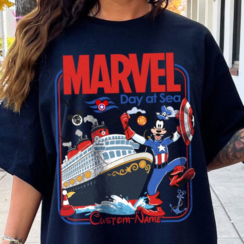 May include: Navy blue t-shirt featuring a graphic of a cruise ship and Mickey Mouse dressed as Captain America. The shirt has the word "MARVEL" in red and "Day at Sea" in white. The shirt also has the text "Custom-Name".