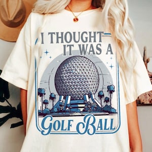 May include: Cream-colored t-shirt with a graphic that reads "I Thought It Was A Golf Ball." The graphic features a geometric sphere design in gray and blue, with a blue border and accents. The shirt is a casual, unisex style.
