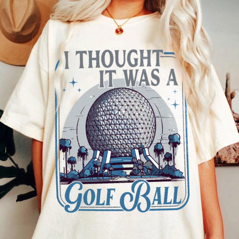 May include: Cream-colored t-shirt with a graphic that reads "I Thought It Was A Golf Ball." The graphic features a geometric sphere design in gray and blue, with a blue border and accents. The shirt is a casual, unisex style.