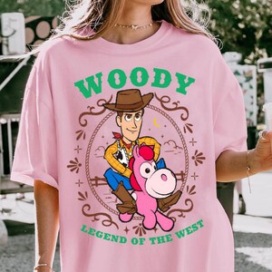 May include: A light pink t-shirt featuring a graphic of the character Woody riding a pink horse. The design includes a brown rope border, a yellow crescent moon, and green text that reads WOODY LEGEND OF THE WEST in a western-style font.