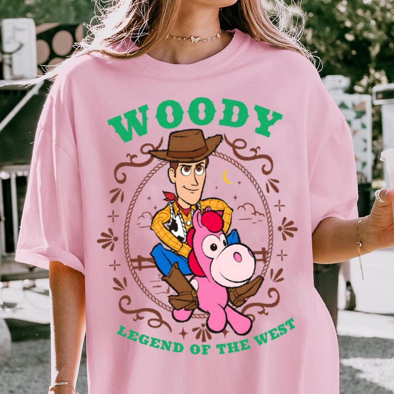 May include: A light pink t-shirt featuring a graphic of the character Woody riding a pink horse. The design includes a brown rope border, a yellow crescent moon, and green text that reads WOODY LEGEND OF THE WEST in a western-style font.