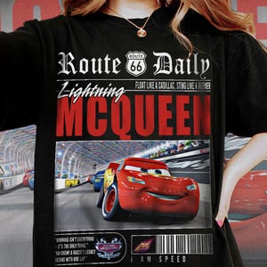 Op de afbeelding: Zwart T-shirt met een afbeelding van Lightning McQueen uit de film Cars. De afbeelding heeft een krantenstijl ontwerp met de tekst "Route 66 Daily" en "Lightning McQueen". De afbeelding bevat ook een citaat uit de film: "Winnen is niet alles - het is het enige." en "Ka-chow! De erfenis van een coureur begint met een ronde."