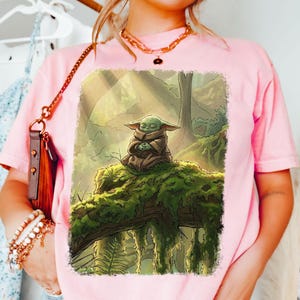 May include: Pale pink t-shirt with a graphic of a green alien character meditating in a forest. The artwork shows the character sitting on a mossy log, with a detailed forest background. The design uses soft colours and shading.