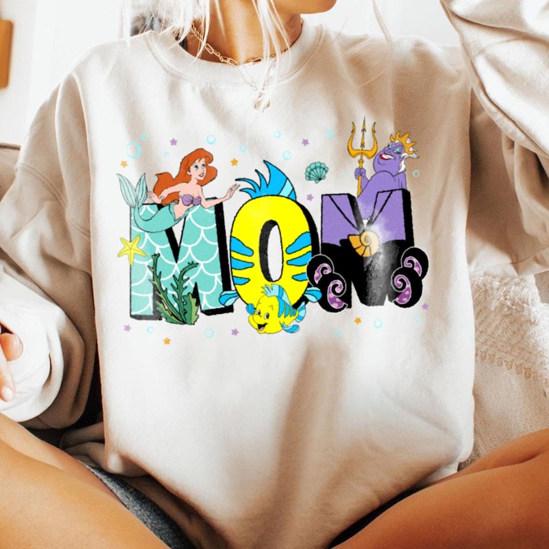 May include: A white sweatshirt with a graphic design of the word "MOM" in colorful letters. The letters are decorated with characters from the Disney movie The Little Mermaid, including Ariel, Flounder, and Ursula.