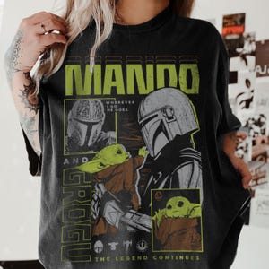 May include: Black t-shirt with a green and yellow graphic design that reads "MANDO". The design includes illustrations of a helmeted character and a small green creature, with the words "Grogu" and "The Legend Continues".