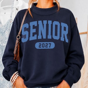 May include: A navy blue long-sleeve sweatshirt with a mock neck collar. The front features the word SENIOR in large, light blue collegiate-style lettering, with the number 2027 printed in light blue inside a rounded rectangular shape below it.