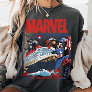 May include: Dark gray sweatshirt featuring a graphic of a cruise ship with Mickey Mouse dressed as Captain America. The text reads "MARVEL Day at Sea" in red and blue, with additional text at the bottom.