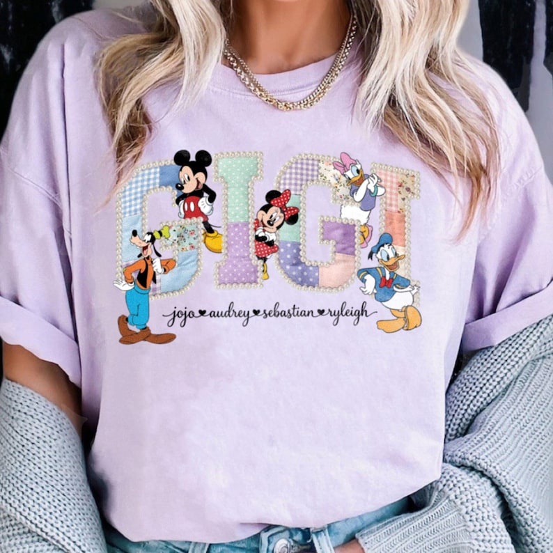 May include: A lilac t-shirt with the word "GIGI" in a patchwork design, featuring Disney characters Mickey Mouse, Minnie Mouse, Donald Duck, Daisy Duck, and Goofy. The names "jojo audrey sebastian ryleigh" are below the word.