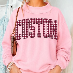 May include: A light pink long-sleeve sweatshirt featuring a mock neck collar. The word CUSTOM is printed across the chest in a dark red and white gingham plaid pattern. A gold chain strap from a bag hangs over the left shoulder of the garment.