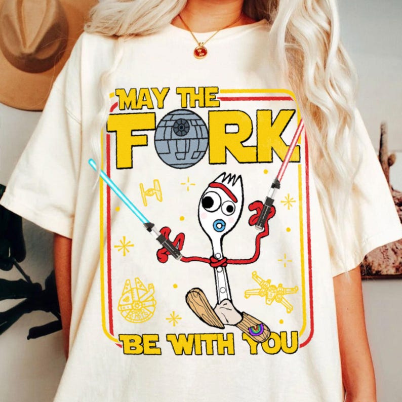 May include: Cream-colored t-shirt with a graphic featuring a spork character holding lightsabers, with the text "MAY THE FORK BE WITH YOU." The design includes a Death Star, starships, and other Star Wars-themed elements. The shirt has a crew neck and short sleeves.