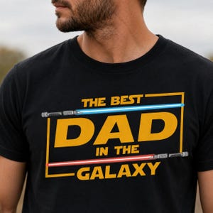 May include: Black t-shirt featuring the text "THE BEST DAD IN THE GALAXY" in yellow, with blue and red lightsaber graphics. The design is centered on the chest. The shirt is made of a soft material.