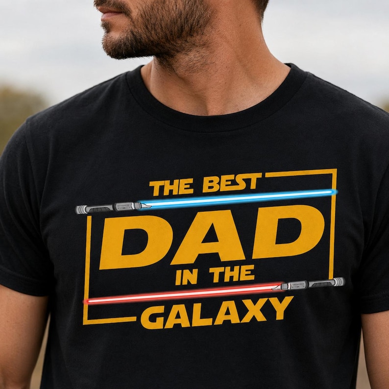 May include: Black t-shirt featuring the text "THE BEST DAD IN THE GALAXY" in yellow, with blue and red lightsaber graphics. The design is centered on the chest. The shirt is made of a soft material.