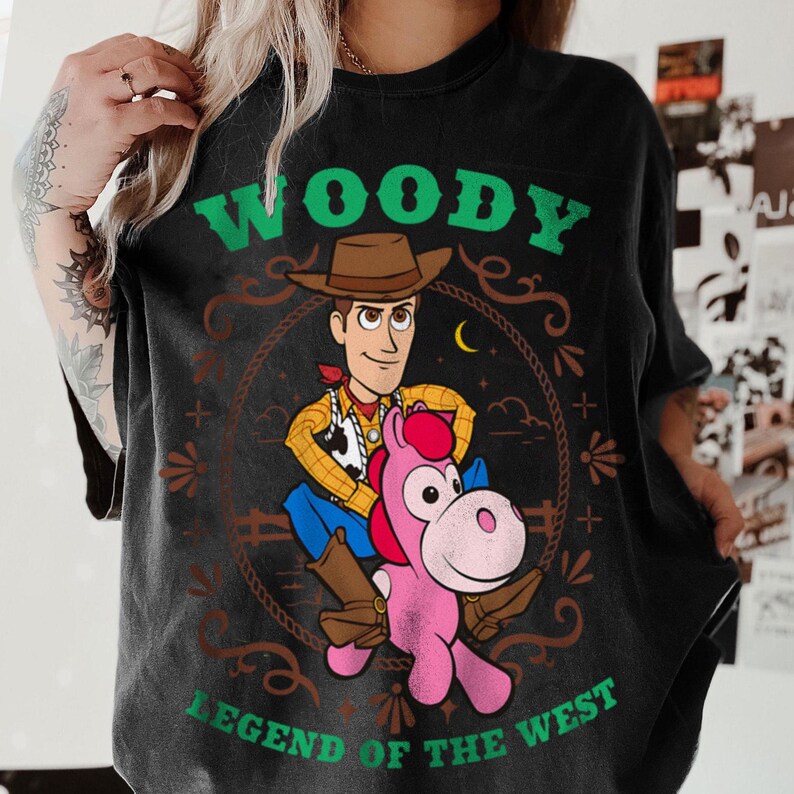 May include: A black t-shirt featuring a graphic print of the character Woody from Toy Story riding a pink horse. The design includes a brown lasso frame, a yellow crescent moon, and the green text Woody Legend of the West printed above and below.
