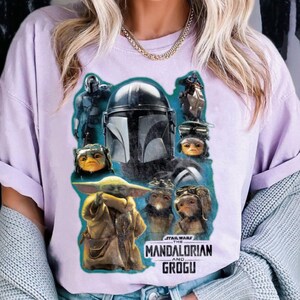 May include: A lilac t-shirt with a graphic print of characters from The Mandalorian and Grogu. The design includes images of the Mandalorian, Grogu, and other alien characters. The Star Wars logo is also visible.