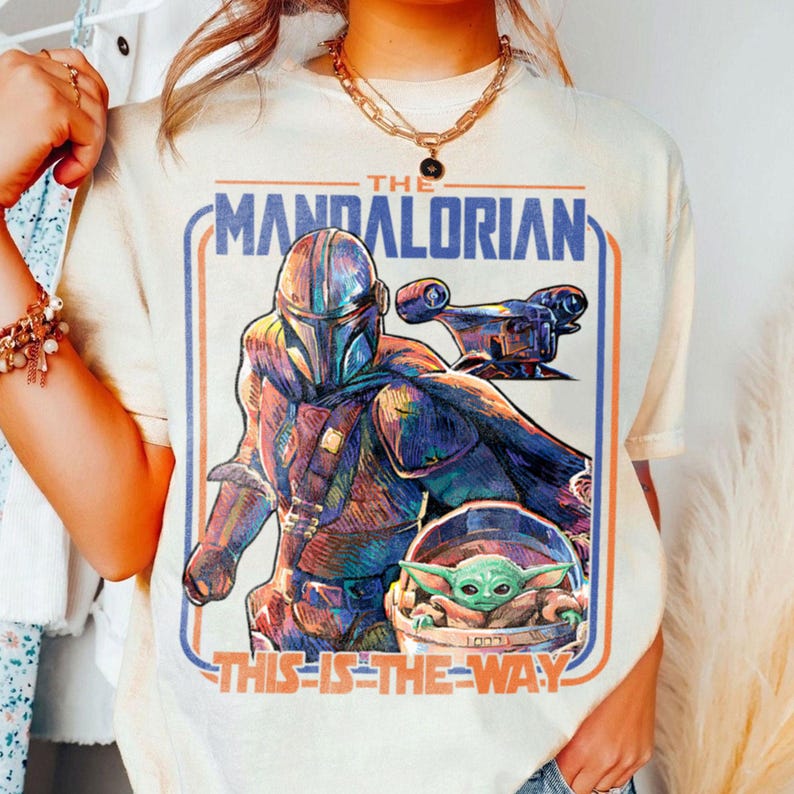 May include: Cream-colored t-shirt featuring a graphic of The Mandalorian, a spaceship, and Grogu. The text "THE MANDALORIAN" and "THIS IS THE WAY" are printed in blue and orange. The design is framed by blue and orange lines.