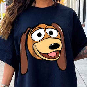 Pu&ograve; includere: T-shirt blu navy con un'illustrazione a cartoni animati di una faccia di cane. Il cane ha grandi occhi, un muso marrone e lunghe orecchie marroni. La bocca del cane &egrave; aperta, rivelando una lingua rosa. Il disegno &egrave; centrato sul davanti.