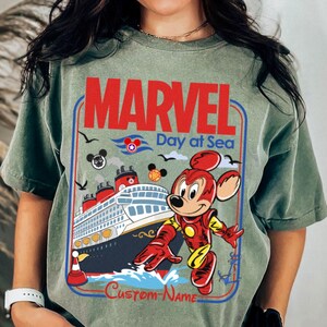 May include: Sage green t-shirt featuring a graphic with the text "MARVEL Day at Sea." The design includes a cruise ship, Mickey Mouse in an Iron Man suit, and other Marvel and Disney-themed elements. The words "Custom-Name" are at the bottom.