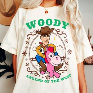 May include: A cream-colored t-shirt featuring a graphic of the character Woody riding a pink horse. The design is enclosed in a rope border with the green text "WOODY" above and "LEGEND OF THE WEST" below, including stars and decorative elements.