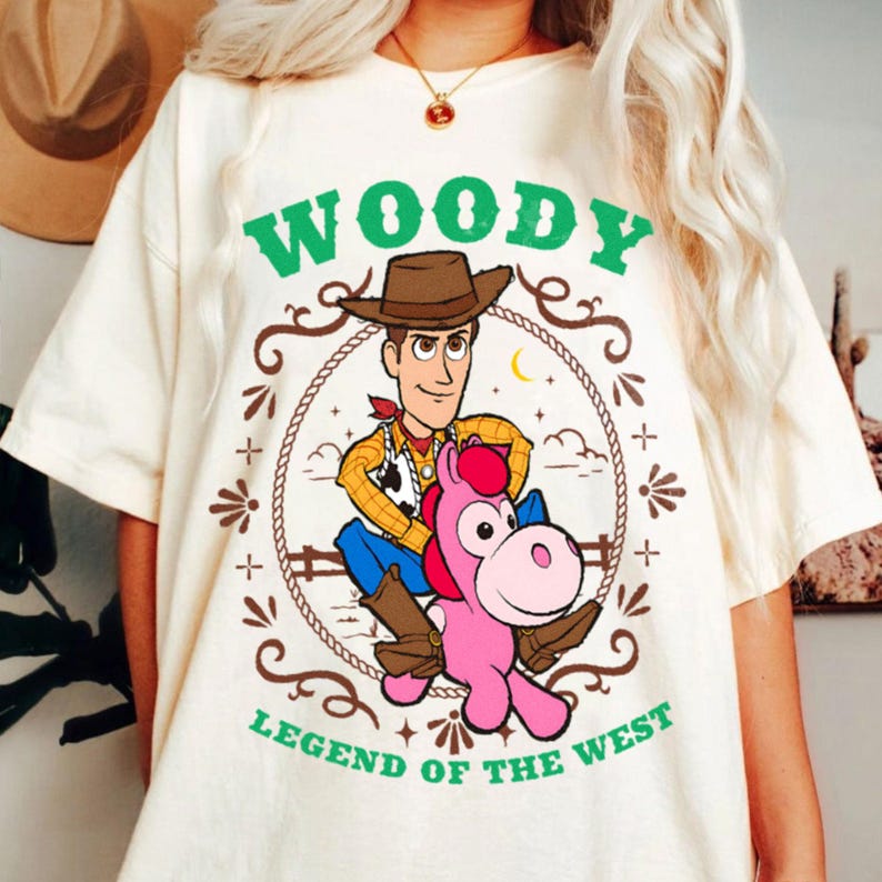 May include: A cream-colored t-shirt featuring a graphic of the character Woody riding a pink horse. The design is enclosed in a rope border with the green text "WOODY" above and "LEGEND OF THE WEST" below, including stars and decorative elements.