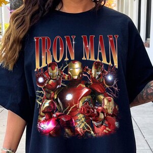 Puede incluir: Camiseta azul marino con un dise&ntilde;o de Iron Man en rojo y dorado. El dise&ntilde;o presenta m&uacute;ltiples figuras de Iron Man y el texto "IRON MAN" en dorado, con un efecto desgastado.