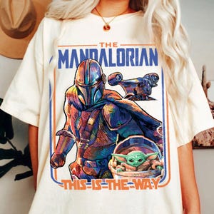 May include: Cream-colored t-shirt featuring a colorful graphic of The Mandalorian, a spaceship, and Grogu. The text "THE MANDALORIAN" is above the image, and "THIS IS THE WAY" is below. The design is framed with blue and orange lines.