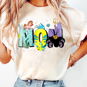 May include: A white t-shirt with a graphic design of the word "MOM" in colorful letters. The letters are decorated with characters from the Little Mermaid, including Ariel, Flounder, and Ursula.