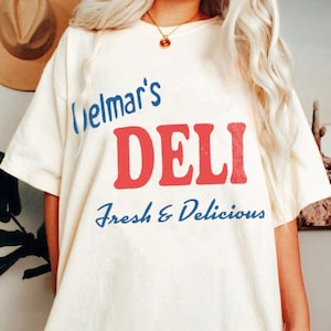 May include: An off-white short-sleeve t-shirt featuring the blue text Delmar's, the large red text DELI, and the blue text Fresh & Delicious. The shirt is worn with a gold chain necklace featuring a small round pendant.